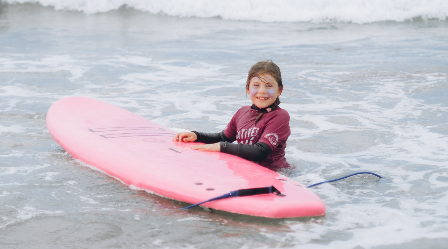 Smiling child in a wetsuit with face sunscreen holding a pink surfboard in shallow ocean water.