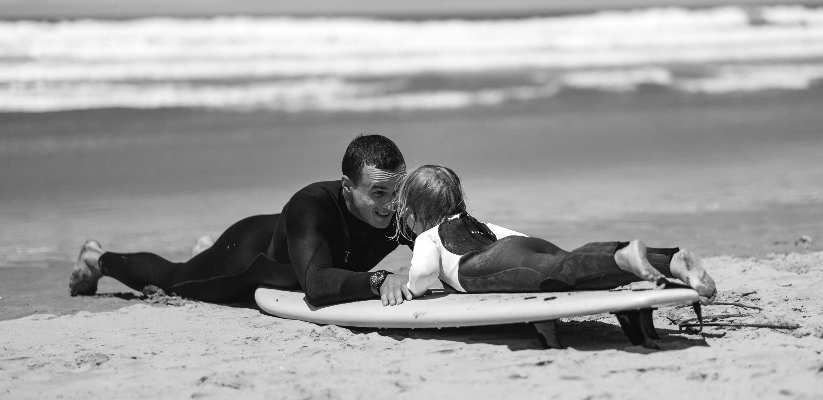 Adult and child lying on a surfboard on the beach, facing each other and smiling.