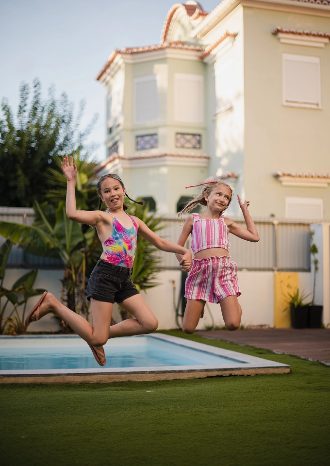 Two girls holding hands and jumping near a backyard pool with a house in the background.