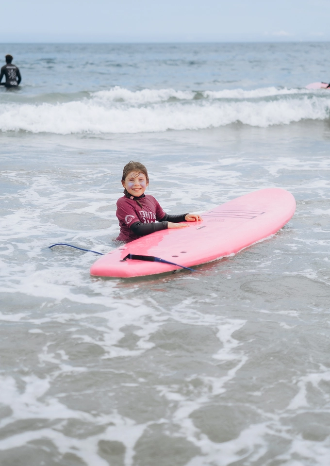 Smiling child in wetsuit holding a pink surfboard in shallow ocean waves.