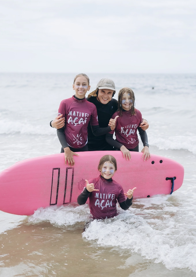 Smiling group of three girls and one man posing with a pink surfboard in shallow ocean water.