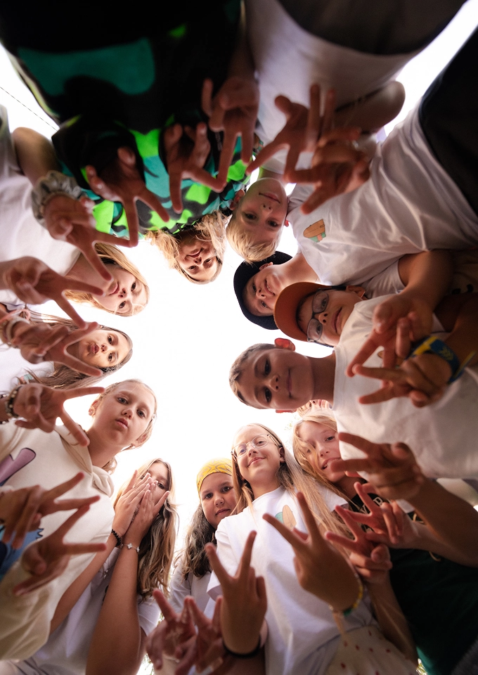 Group of children in a circle looking down at the camera, making peace signs with their hands.