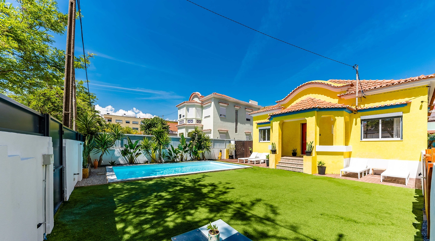 Backyard of yellow house with swimming pool, green lawn, lounge chairs, and potted plants under clear blue sky.