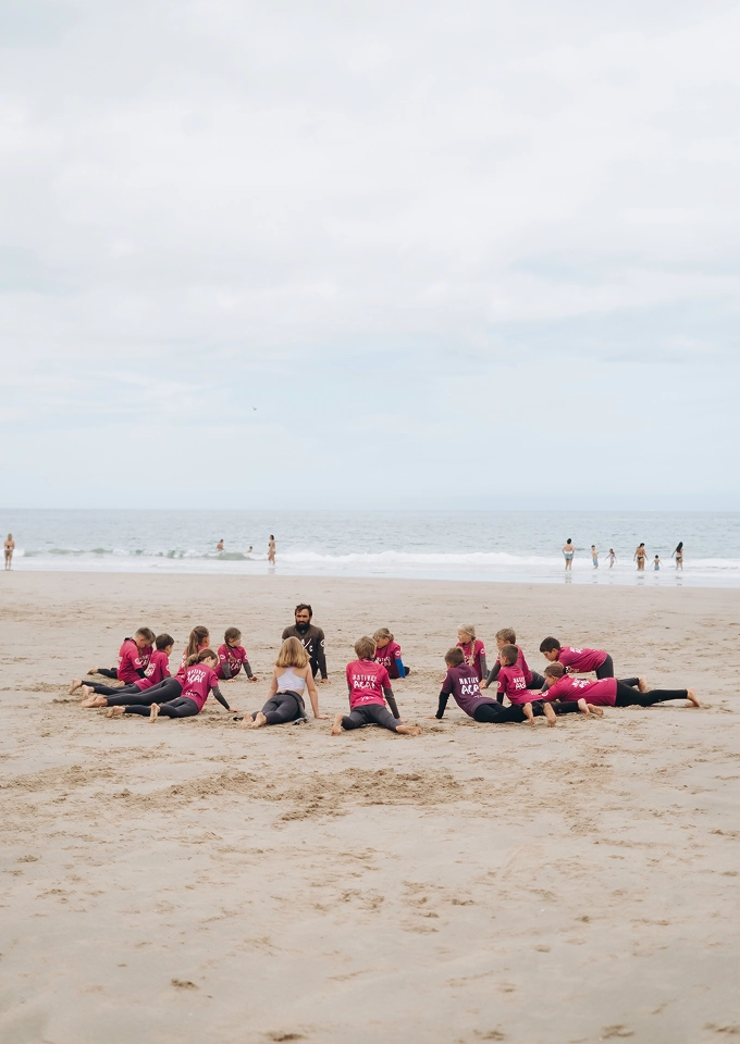 Group of children in pink shirts sitting on sand in a circle around an instructor on a beach.
