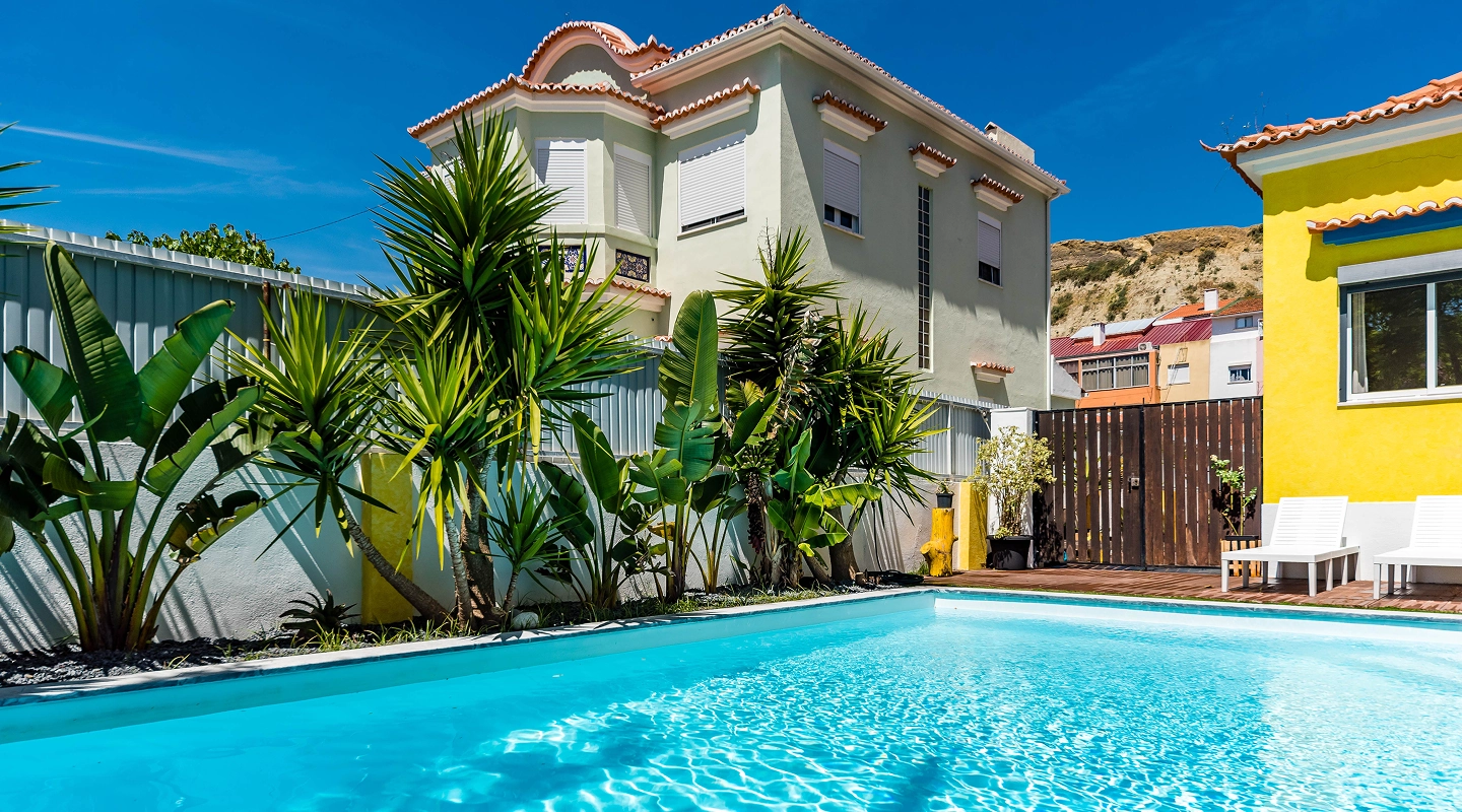 Bright blue swimming pool beside a house with tropical plants and yellow wall under a clear sky.