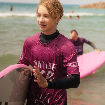 Teen surfer wearing a maroon shirt holding a pink surfboard near the shore.