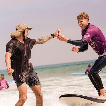 Two men smiling and giving a high-five while surfing in the ocean near the shore.