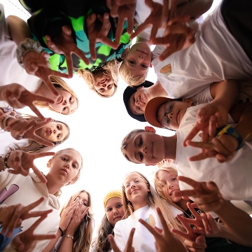 Group of children forming a circle and making peace signs while looking down at the camera.