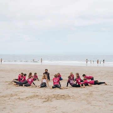 Group of children in matching pink shirts sitting in a circle on a sandy beach with calm sea in the background.